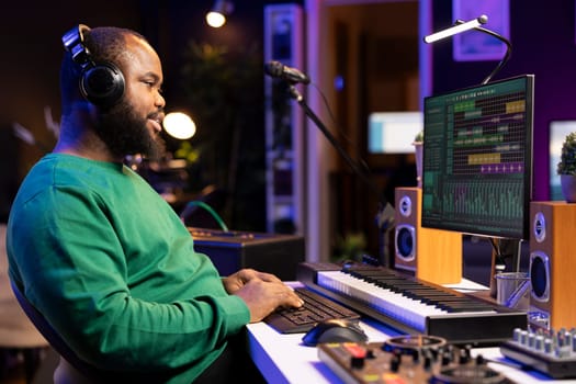 African american composer mixing and adding sounds on his recordings, listening to the final product on headset. Audio engineer producing new music, editing volume levels in home studio.