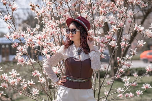 Magnolia park woman. Stylish woman in a hat stands near the magnolia bush in the park. Dressed in white corset pants and posing for the camera