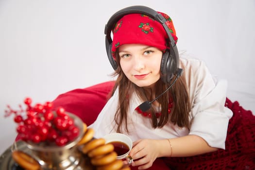 Young girl in red scarf and large headphones with microphone with samovar, bagels, cap of tea on Easter or Maslenitsa. Woman radio or television presenter, female telecom operator, blogger, Freelancer