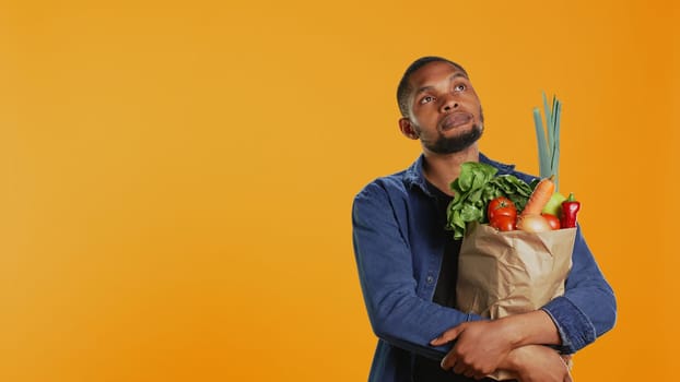 Male model thinking about new vegan food recipe to cook with his freshly harvested homegrown produce in a paper bag. Young guy enjoys healthy eating, organic nutrition. Camera B.