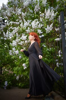 Elegant senior mature Woman in Black Dress by Blooming Lilac Bush at Dusk. Woman with red hair stands poised among lilac blooms