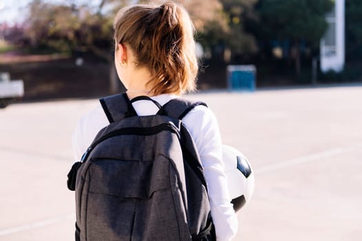 rear view of an unrecognizable young caucasian woman with backpack in a urban football court with a soccer ball in hand, concept of sport and active lifestyle