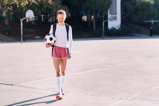 smiling young caucasian woman with backpack walking at urban football court with a soccer ball in hand, concept of sport and active lifestyle, copy space for text