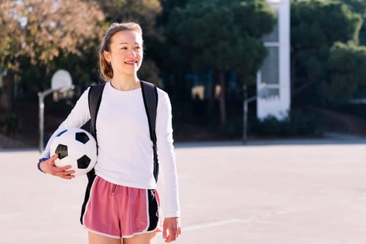 smiling young caucasian woman with backpack walking at urban football court with a soccer ball in hand, concept of sport and active lifestyle, copy space for text