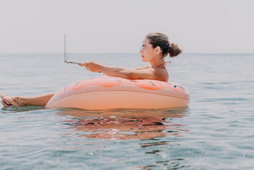 Woman freelancer works on laptop swimming in sea on pink inflatable ring. Pretty lady typing on computer while floating in the sea on inflatable donut at sunset. Freelance, remote work on vacation