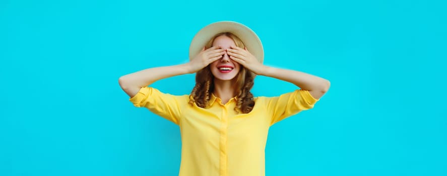 Stylish surprised happy young woman covering her eyes with hands wearing summer straw hat, yellow shirt on blue background