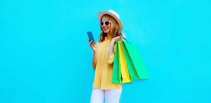 Beautiful happy young woman looking at mobile phone with shopping bag, wearing summer hat on blue studio background