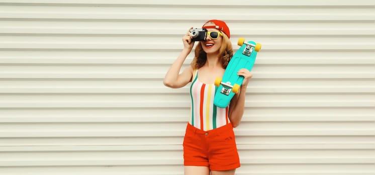 Summer portrait of happy cheerful stylish young woman with skateboard takes picture with camera in colorful clothes on white background