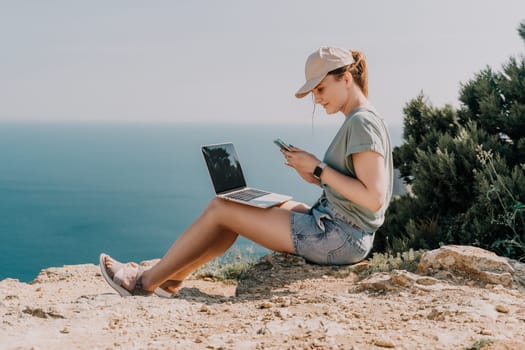 Digital nomad, woman in the hat, a business woman with a laptop sits on the rocks by the sea during sunset, makes a business transaction online from a distance. Freelance, remote work on vacation.