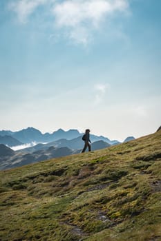 A person is walking up a grassy hill in the mountains under a cloudy sky, surrounded by lush greenery with a vast landscape spreading in the horizon