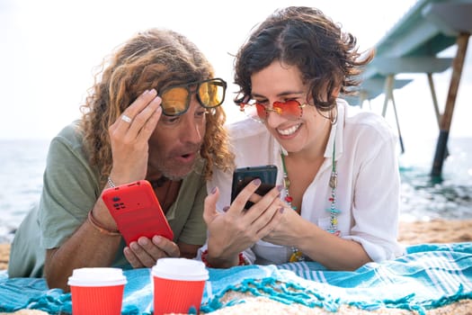 Middle-aged man and woman sitting on the beach with surprised face, browsing smartphone apps. Concept:Holidays y Technology