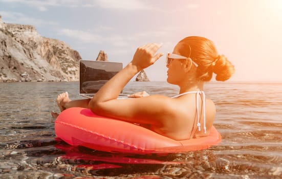 Woman freelancer works on laptop swimming in sea on pink inflatable ring. Pretty lady typing on computer while floating in the sea on inflatable donut at sunset. Freelance, remote work on vacation