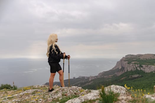 A woman stands on a hill overlooking the ocean. She is wearing a black jacket and shorts. The sky is cloudy and the ocean is in the background