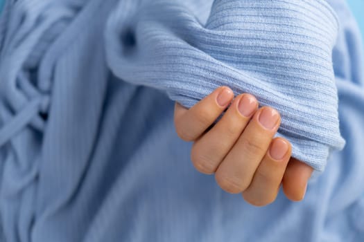 Woman manicured hands, stylish beige nails. Closeup of manicured nails of female hand in blue sweater in blue background. Winter or autumn style of nail design concept. Beauty treatment.