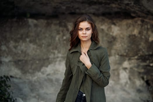 Confident woman in green jacket and jeans standing in front of rugged rock wall with hands on hips