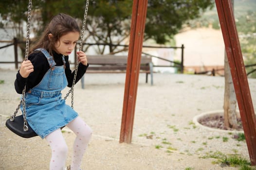 Adorable Caucasian little child girl in denim sundress, swinging on the outdoors playground. People. Happy carefree childhood concept. Lifestyle