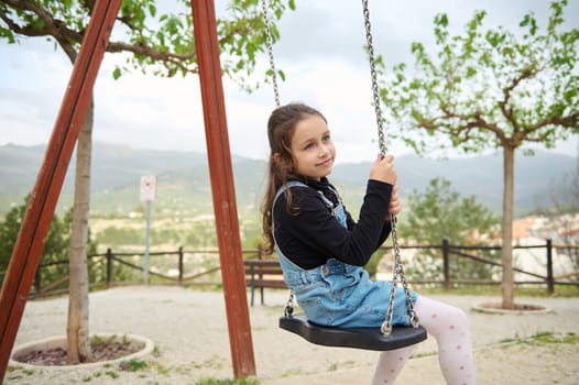 Little child girl 6 years old smiling and dreamily looking away while sitting on a wooden swing in the playground outdoors. People. Happy carefree childhood. Recreation