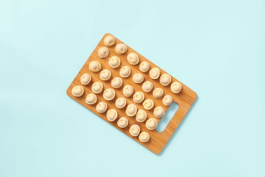 Top view of a wooden board with neatly laid out semi-cooked dumplings on blue background.