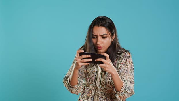 Gamer with disappointed look on face after receiving game over screen, holding smartphone. Indian woman upset after losing videogame, playing with phone, isolated over studio background