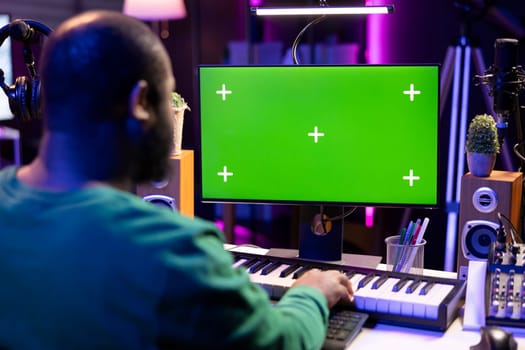 Producer composing music with electronic keyboard notes and greenscreen display on pc, post production in home studio. Musician artist plays midi controller next to mockup layout.