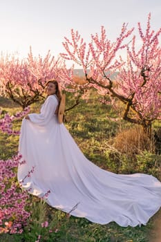 Woman blooming peach orchard. Against the backdrop of a picturesque peach orchard, a woman in a long white dress and hat enjoys a peaceful walk in the park, surrounded by the beauty of nature