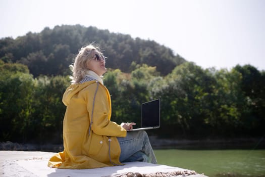A woman in a yellow coat is sitting on a dock with a laptop open in front of her. She is enjoying the view of the water and the surrounding nature