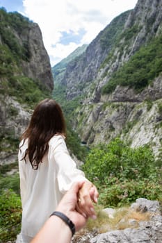 A girl stands with her back turned and a man holds her hand against the backdrop of a mountain view with lush green rocks under a cloudy sky