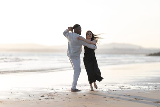 A man and a woman are dancing on the beach. The man is wearing a white shirt and the woman is wearing a black dress. Scene is joyful and carefree