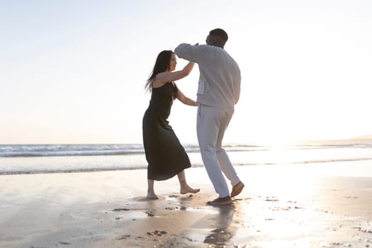 A man and woman are dancing on the beach. The man is wearing a white shirt and the woman is wearing a black dress. The sun is setting in the background, creating a warm and romantic atmosphere