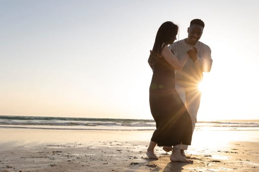 A couple dancing on the beach in the sun. The man is wearing a white shirt and the woman is wearing a black dress