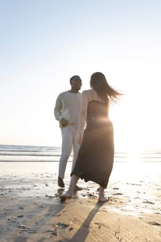 A man and woman are standing on the beach, smiling at each other. The woman is wearing a black dress and the man is wearing a white shirt. The sun is shining brightly, creating a warm
