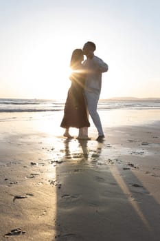 A couple is kissing on the beach at sunset. The woman is wearing a black dress and the man is wearing white pants