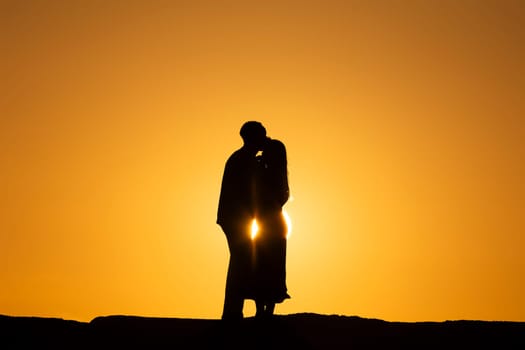 A couple is kissing in front of a sunset. The sun is setting behind them, casting a warm glow on their faces. Concept of intimacy and romance, as the couple shares a tender moment together