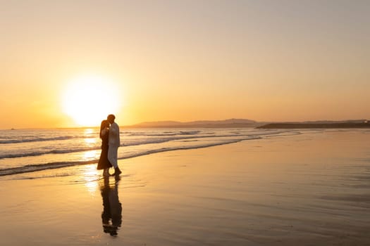 A couple is walking on the beach at sunset. The man is wearing a white shirt and the woman is wearing a black dress. The sun is setting in the background, casting a warm glow over the scene