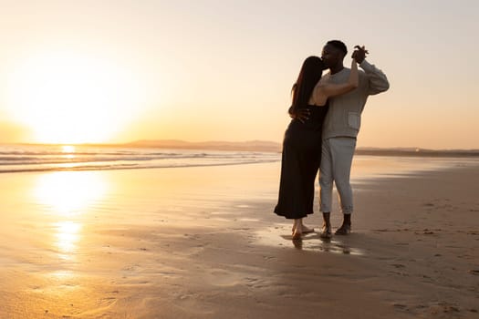 A couple is dancing on the beach at sunset. The man is wearing a sweater and the woman is wearing a black dress