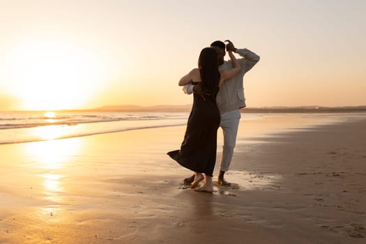 A couple is dancing on the beach at sunset. The man is looking at the woman, and they are both smiling. Scene is romantic and joyful
