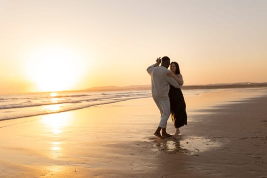 A man and woman are dancing on the beach at sunset. The man is wearing a white shirt and the woman is wearing a black dress. The scene is romantic and peaceful, with the sun setting in the background