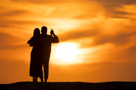 A couple is standing on a hillside, looking at the sun. The sun is setting, and the sky is filled with clouds. Scene is romantic and peaceful