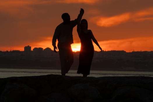 A couple dancing in the sunset. The man is holding the woman's hand. The woman is wearing a black dress