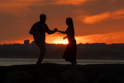 A couple dancing on a beach at sunset. The man is wearing a white shirt and the woman is wearing a black dress. The sky is orange and the water is calm