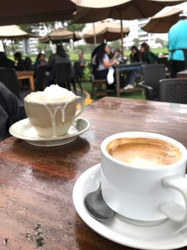 Two cups of coffee on a table at an al fresco cafe with patrons in the background