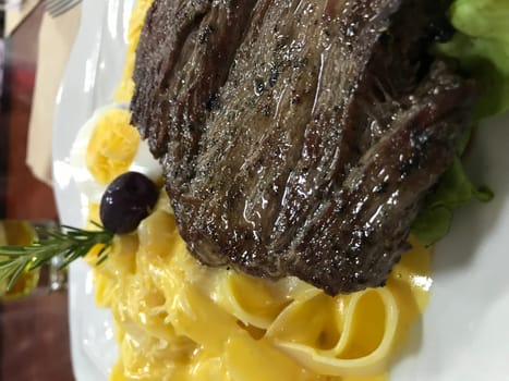 Delectable grilled beef steak served on a bed of creamy pasta, garnished with salad greens, egg, and an olive