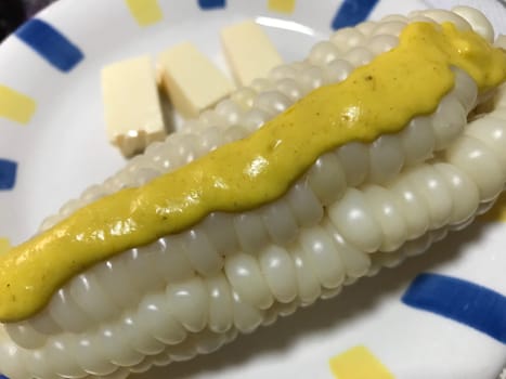 Close-up of a juicy white corn cob smothered with mustard alongside cheese cubes on a plate