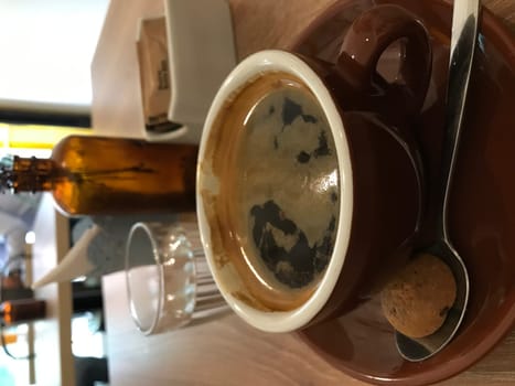 Casual and cozy rustic tabletop coffee scene with brewed espresso in a brown cup and saucer. Accompanied by a spoon. Empty bottle. And morning refreshment on a wooden table