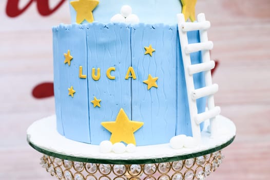 Close-up of a blue fondant birthday cake with the name 'luca', adorned with yellow stars and a white ladder