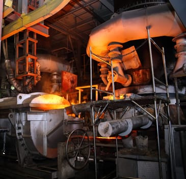 Molten metal is being poured from a ladle into a mold in a steel mill. The factory is filled with heavy industry machinery and equipment. This is a metallurgy and metalworking theme.
