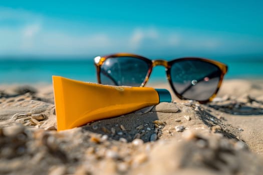 A yellow sunscreen tube and a pair of sunglasses lie on a sandy beach.