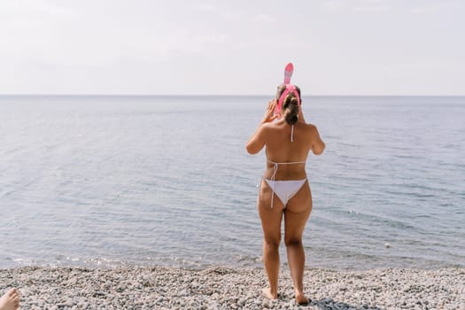 A woman in a bikini stands on a beach, looking out at the ocean. She is holding a pink snorkel and she is enjoying the view