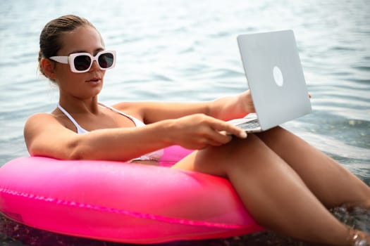 A woman is sitting on a pink inflatable raft with a laptop in her lap. She is wearing sunglasses and a white bikini top. Concept of relaxation and leisure