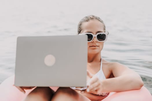 A woman is sitting on a pink inflatable raft with a laptop in her lap. She is wearing sunglasses and she is enjoying the sun and water